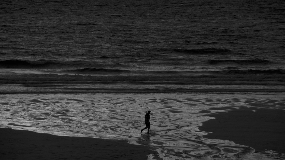 A black and white image of a man walking along the shoreline of a beach, his silhouette outlined in the middle of the frame.