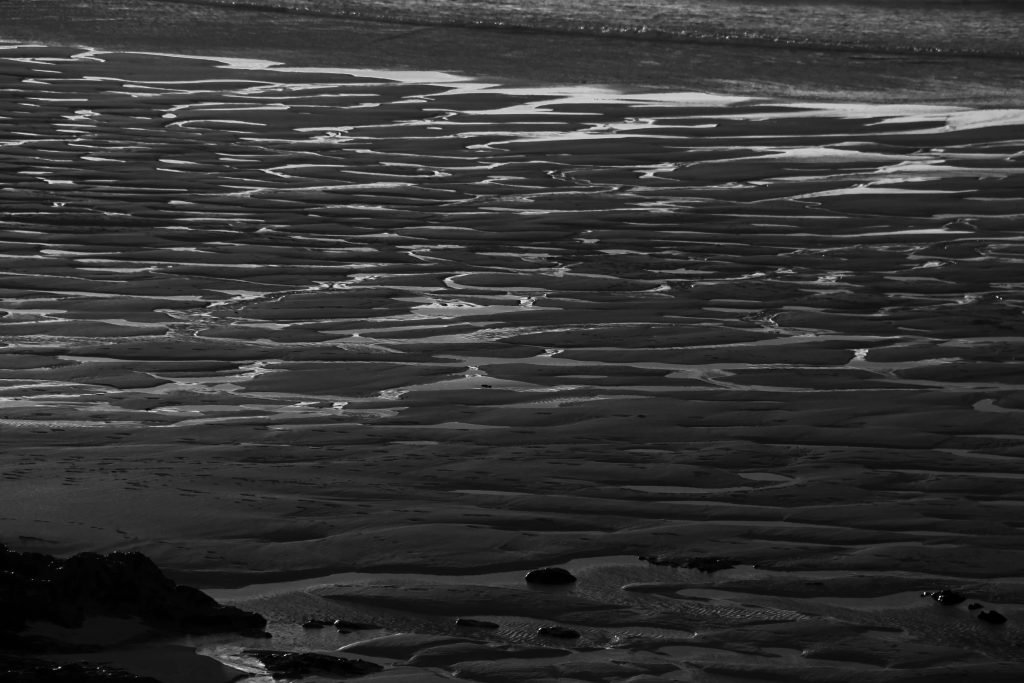 A black and white shoreline at a beach, glistening water trails reflecting the light.