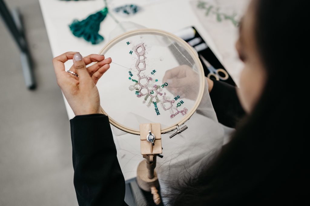 A person hand-embroidering a chemical structure which is placed in an embroidery hoop. 