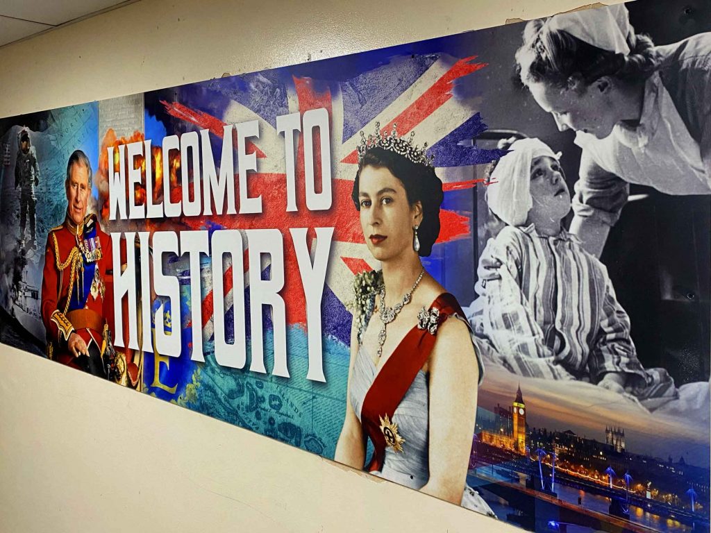 wall graphic with a large text saying welcome to history with a Union Jack behind with Queen Elizabeth and King Charles with the moon landing and and black and white picture of a nurse and a child