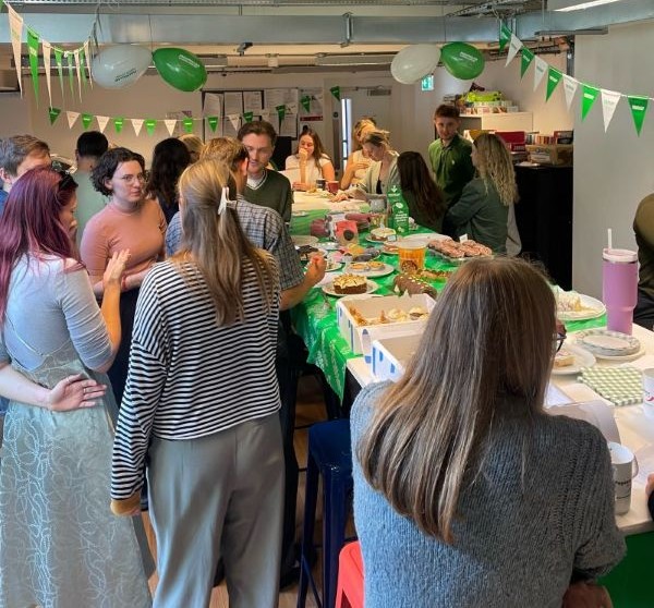Group of people chatting with green and white decorations. They are surrounding a table with cakes and sweets. 